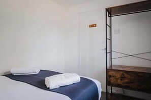 Ett hotellrum med en säng, två handdukar, en hylla i trä och en dörr med ett handtag.