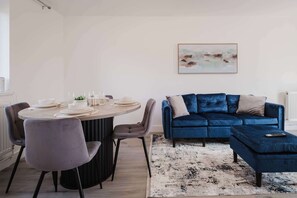 A modern living room with a round dining table set for four, a blue velvet sofa, and a contemporary abstract painting on the wall. - Modern 3 Bedroom House With WiFi (Northampton)