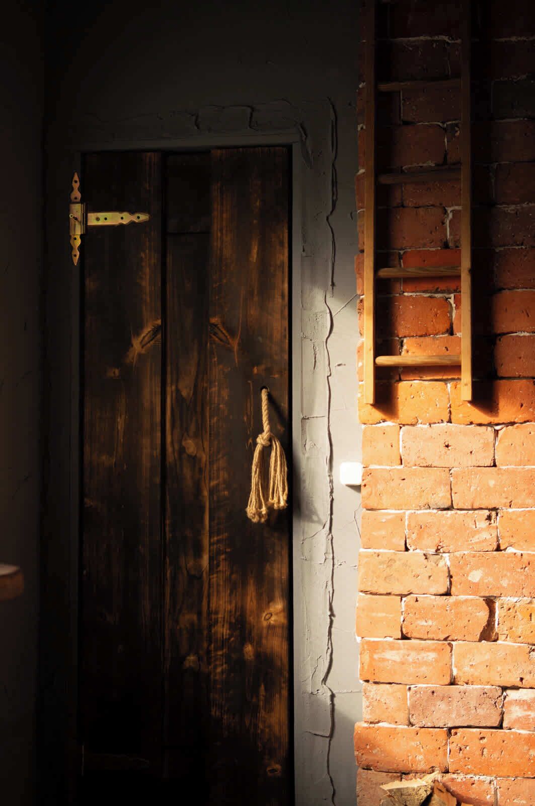 Una porta de fusta amb una tranca de metall i una corda lligada, una paret de maó amb una prestatgeria de fusta, i una vista parcial d'una habitació amb una paret blanca i una finestra.