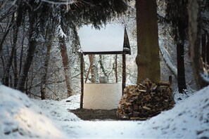 A snow-covered outdoor area with a wooden structure, a stack of firewood, and a stone wall.