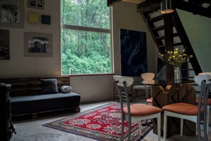 A room with a large window, a sofa, a dining table with chairs, and a red patterned rug.