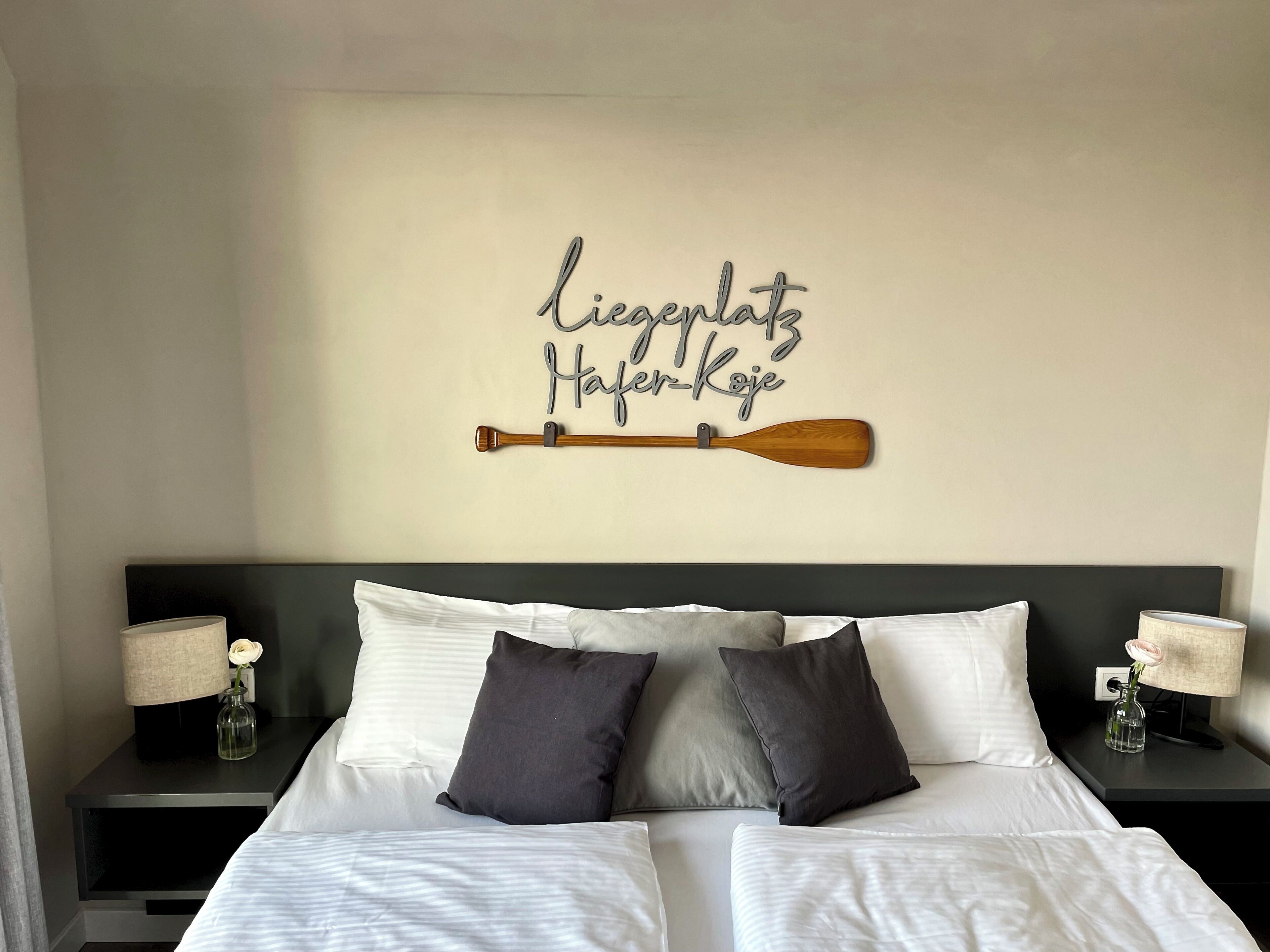Una cama bien hecha con sábanas blancas y dos almohadas, un remo de madera sujeto a la pared y un cartel con texto.