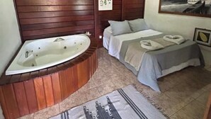 Een kamer met een houten plafond, een ruime badkuip, een bed met een grijze dekbedovertrek en een vloerkleed met een geometrisch patroon.