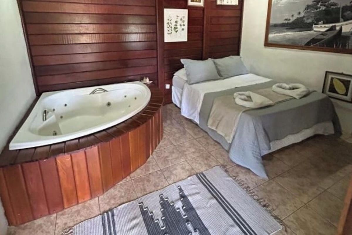 Quarto com teto de madeira, uma banheira grande, cama com colcha cinza e tapete com padrão geométrico.