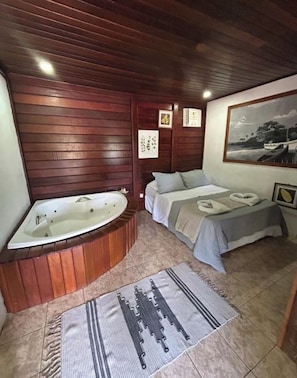 Quarto com teto de madeira, uma banheira grande, cama com colcha cinza e tapete com padrão geométrico.
