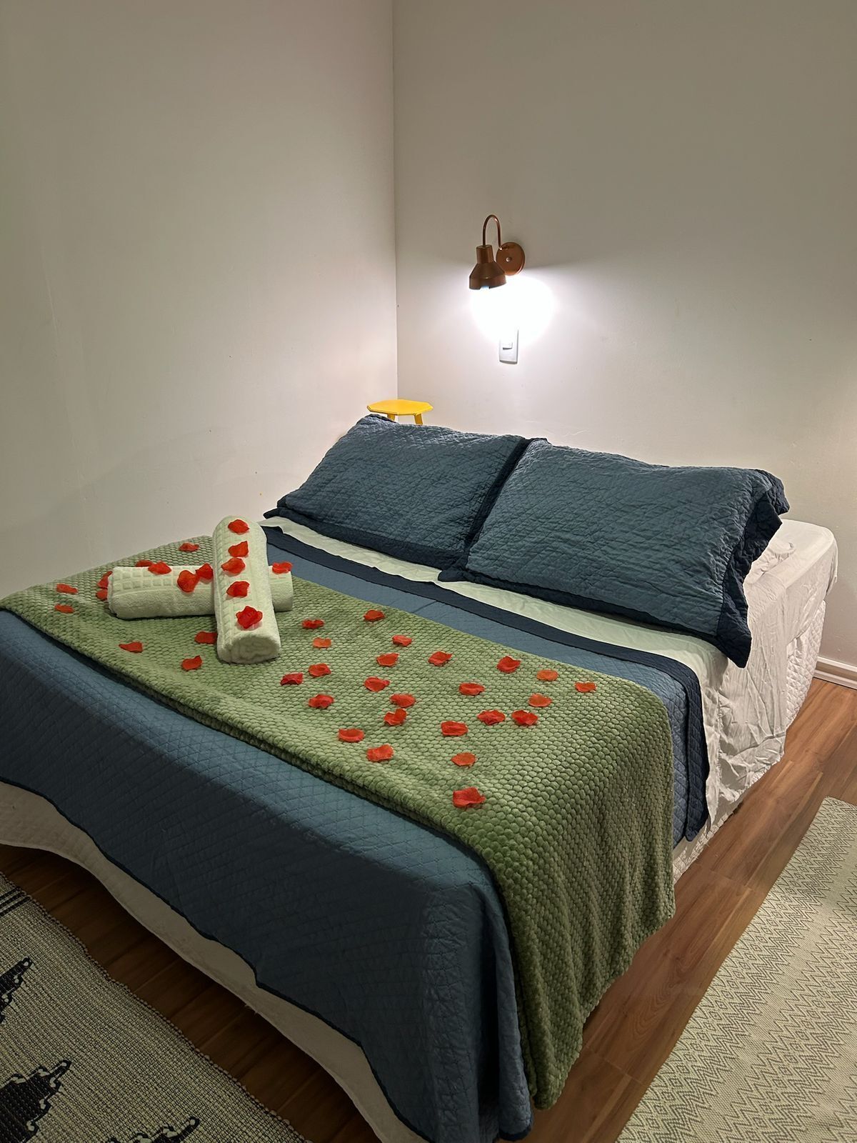 Cama bem arrumada com colcha azul e verde, colchão branco e um banquinho amarelo encostado na parede.