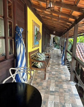 A covered outdoor seating area with wooden benches and chairs, a tiled floor, and a yellow wall with a decorative piece.