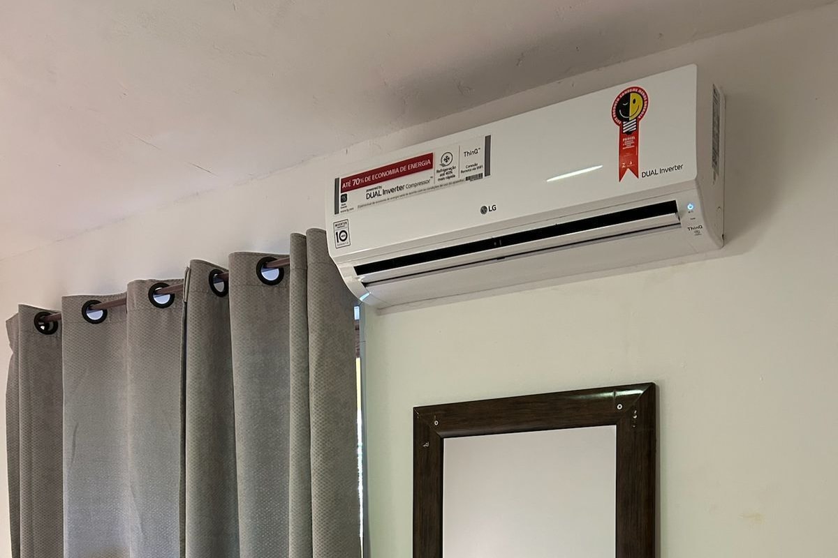 Quarto com ar-condicionado fixado na parede, cortinas cinzas e um espelho refletindo uma cama com cobertor azul.