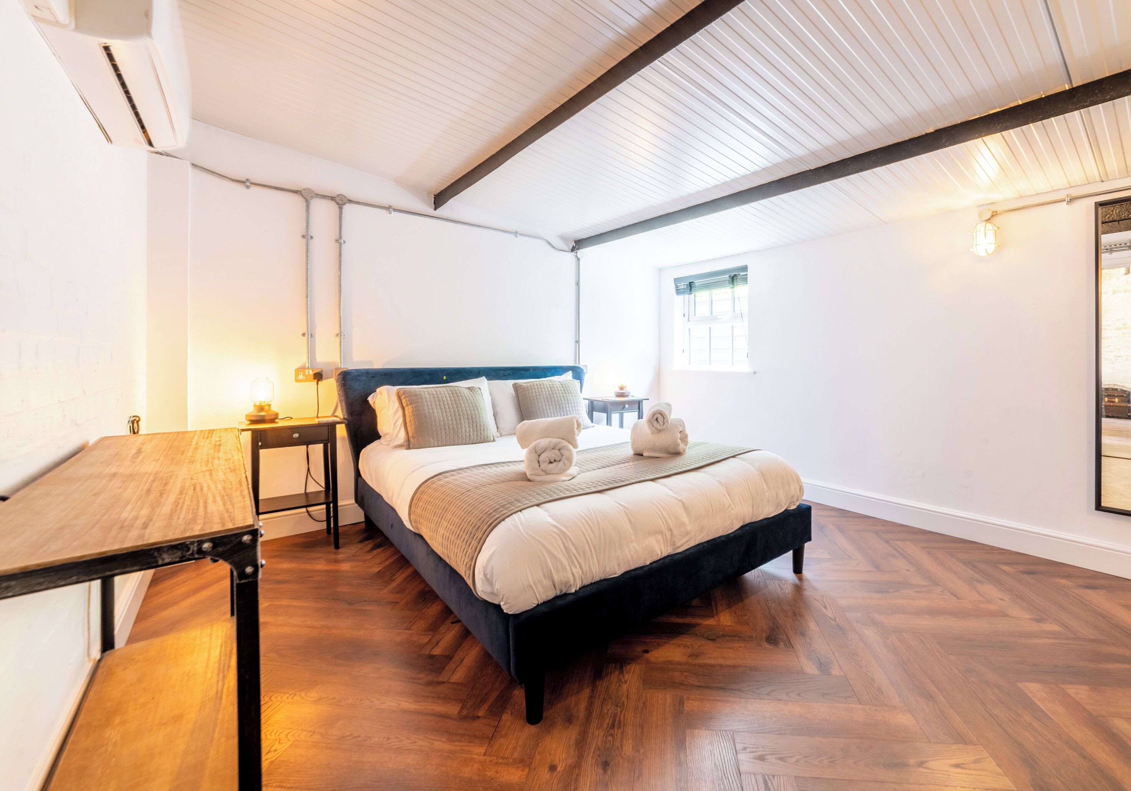 A bedroom with a bed, a nightstand, a table, and a window.