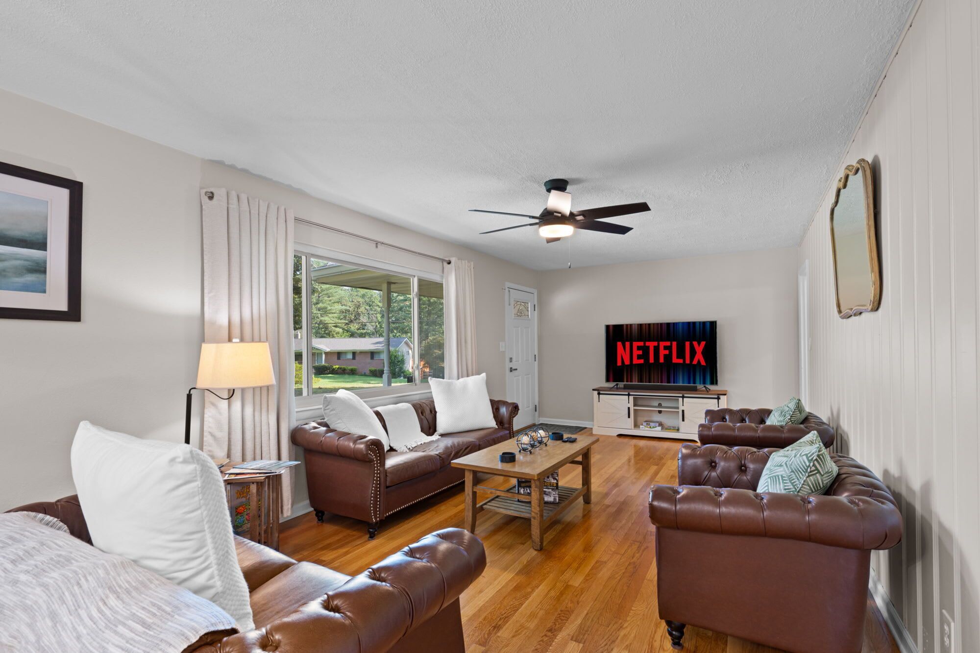 A living room with leather sofas, a coffee table, a ceiling fan, a TV displaying 'NETFLIX', and a window with curtains.