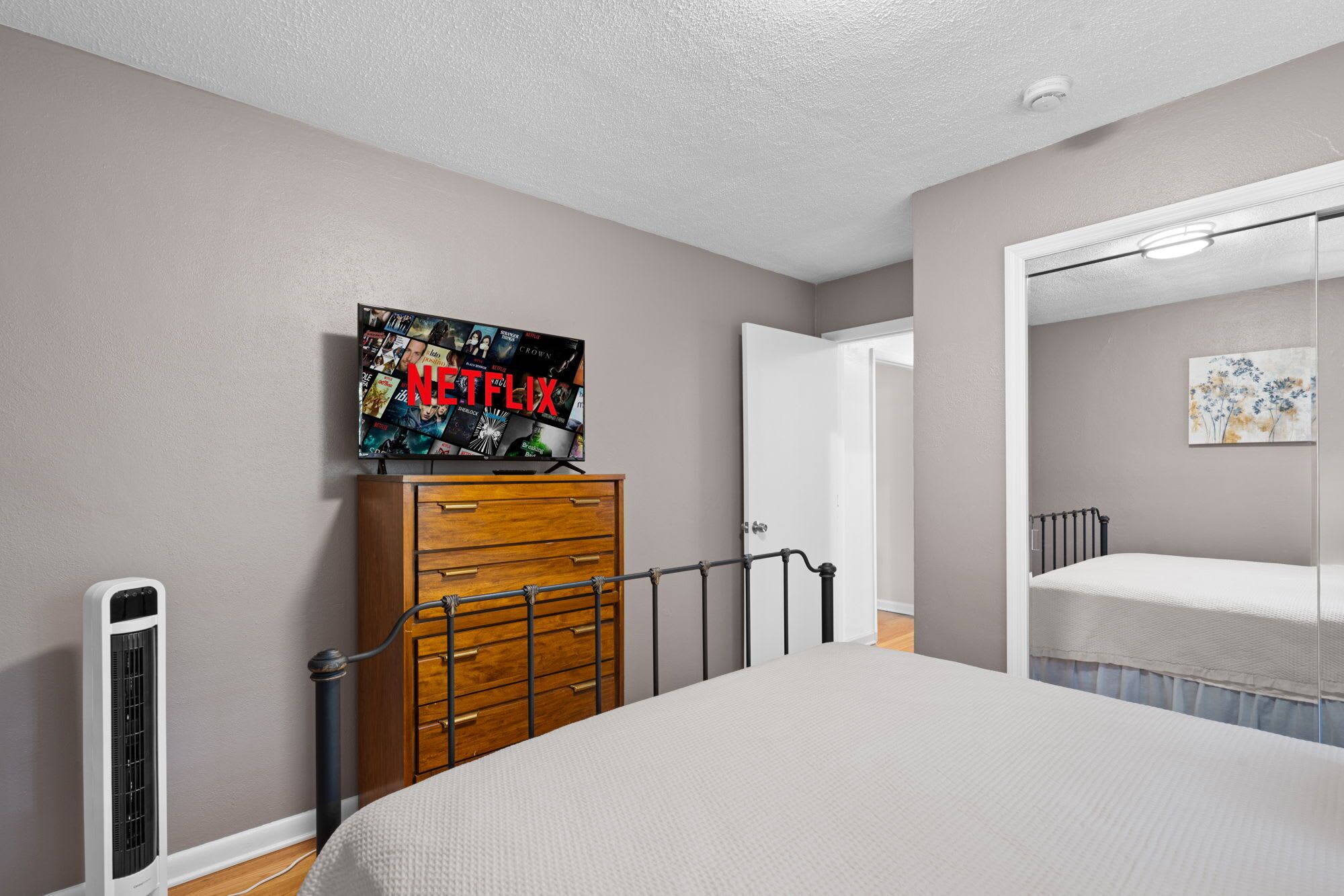 Un dormitorio con cama, cómoda, televisor mostrando Netflix y puerta que da a otra habitación.