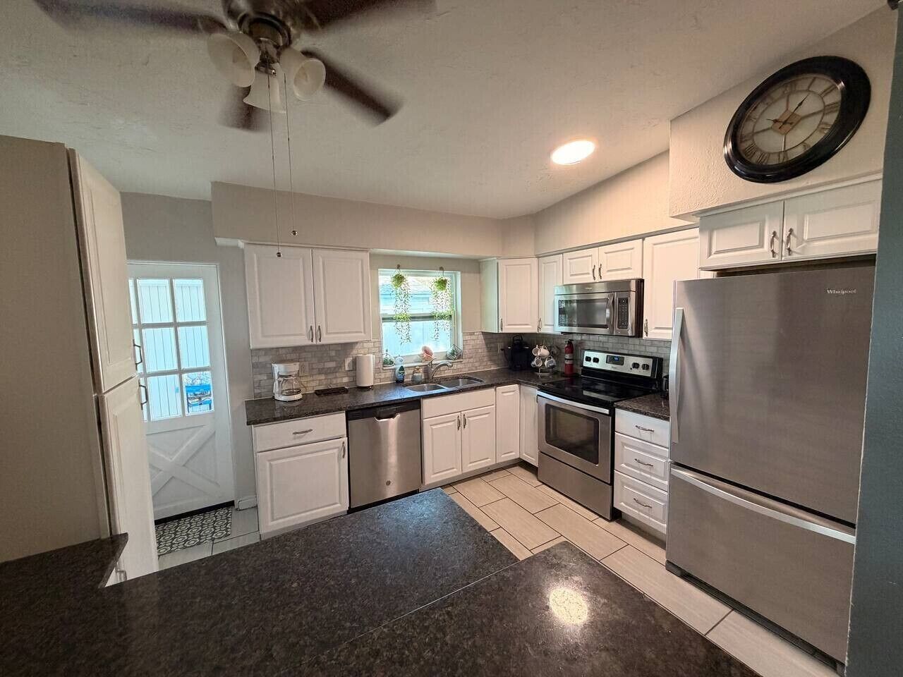 A kitchen with white cabinets, stainless steel appliances, a clock on the wall, and a ceiling fan.
