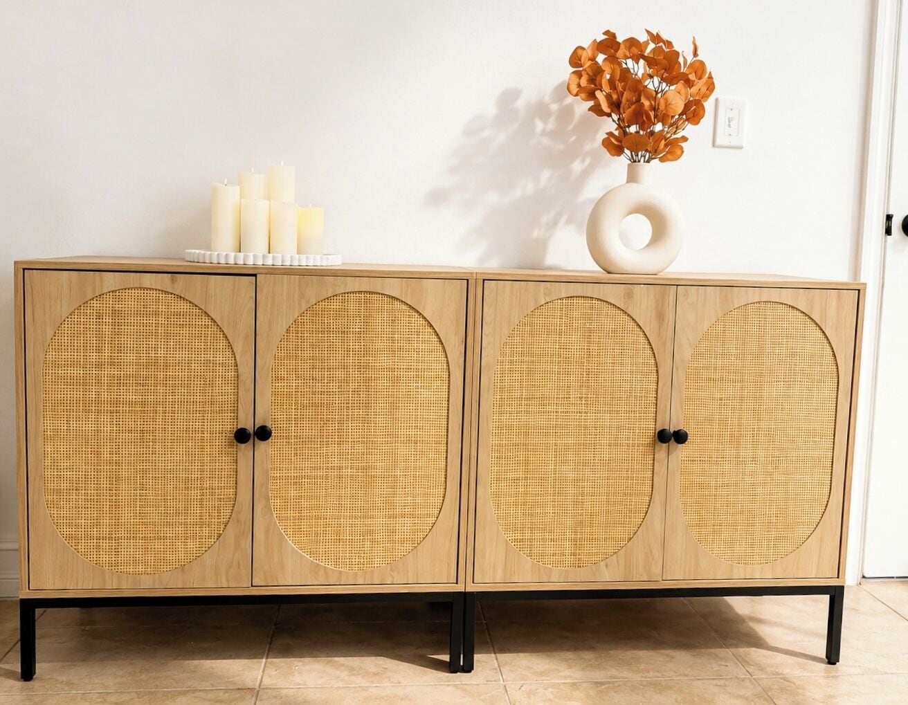 A wooden cabinet with circular woven doors, a vase with dried flowers, and candles on top.