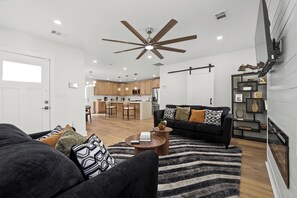 A modern living room with a black sofa, a coffee table, and a striped rug. There is a kitchen in the background with wooden cabinets and a bar area.