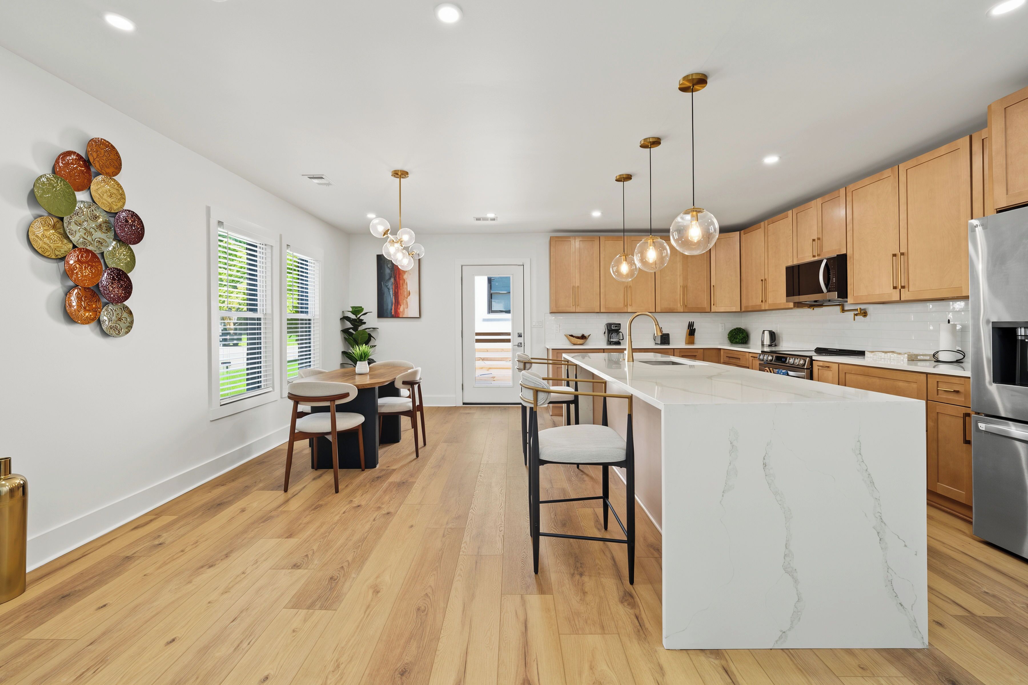 A modern kitchen with wooden cabinets, a central island, pendant lighting, and a dining area with a table and chairs.