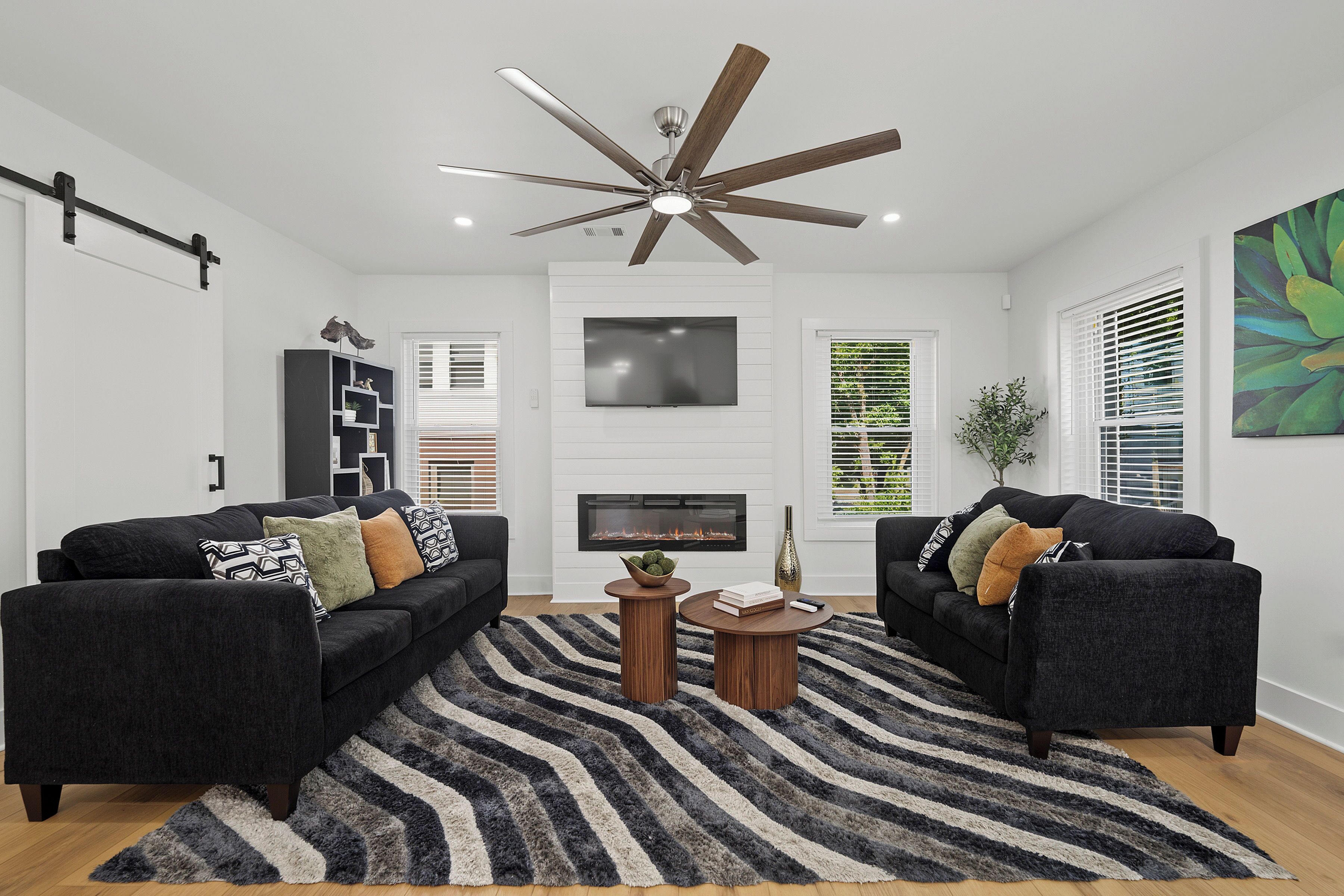Un salon moderne avec deux canapés, une cheminée, un ventilateur de plafond et une grande fenêtre équipée de stores.