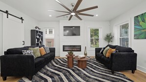 A modern living room with two sofas, a fireplace, a ceiling fan, and a large window with blinds.