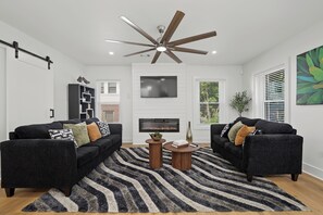 A modern living room with two sofas, a fireplace, a ceiling fan, and a large window with blinds.