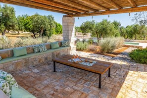 An outdoor patio with a wooden table, stone seating, and a view of trees and a building. - Villa Simeone by Villa Plus (Ostuni)