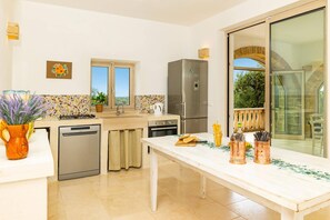A modern kitchen with a dishwasher, stove, and refrigerator. There is a dining table with cutlery and a vase of flowers. - Villa Simeone by Villa Plus (Ostuni)