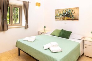 A bedroom with a bed, two pillows, a nightstand with a lamp, a painting on the wall, and a window with curtains. - Villa Simeone by Villa Plus (Ostuni)