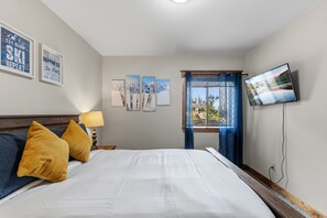 Ein Schlafzimmer mit Bett, Kissen, Nachttisch, Lampe, Fernseher und einem Fenster mit Aussicht.