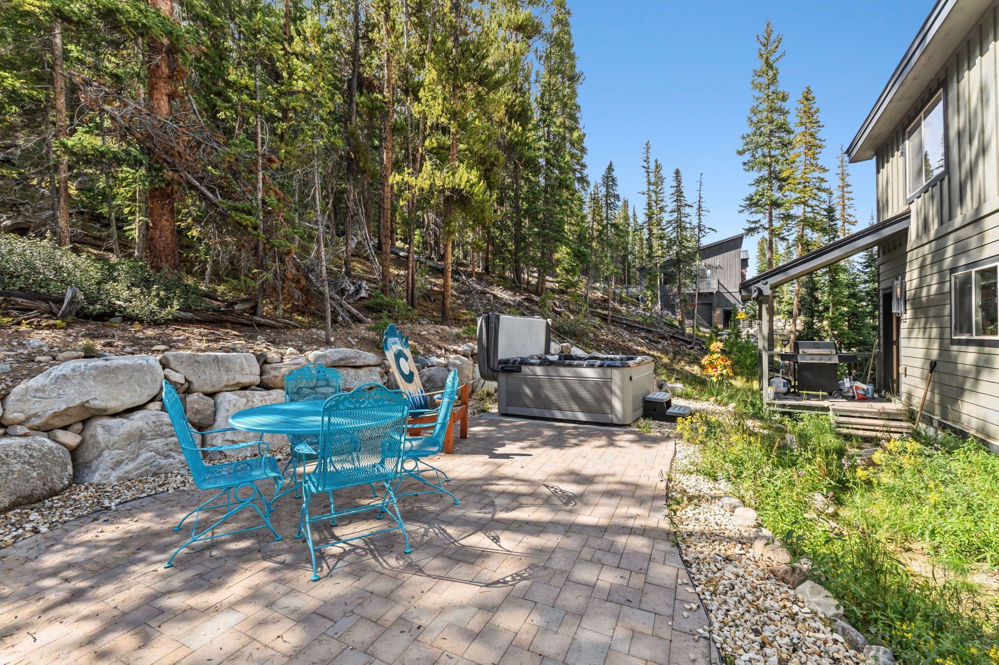 A patio with a table and chairs, a grill, and a house in a forested area.