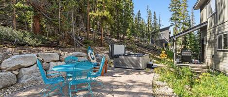 A patio with a table and chairs, a grill, and a house in a forested area.