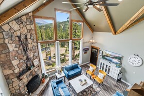 A spacious living area with a stone fireplace, large windows offering mountain views, a ceiling fan, and a clock on the wall. - Scenic and Spacious 4 Bedroom, 3.5 Bath Just Outside of Breckenridge (Blue River)