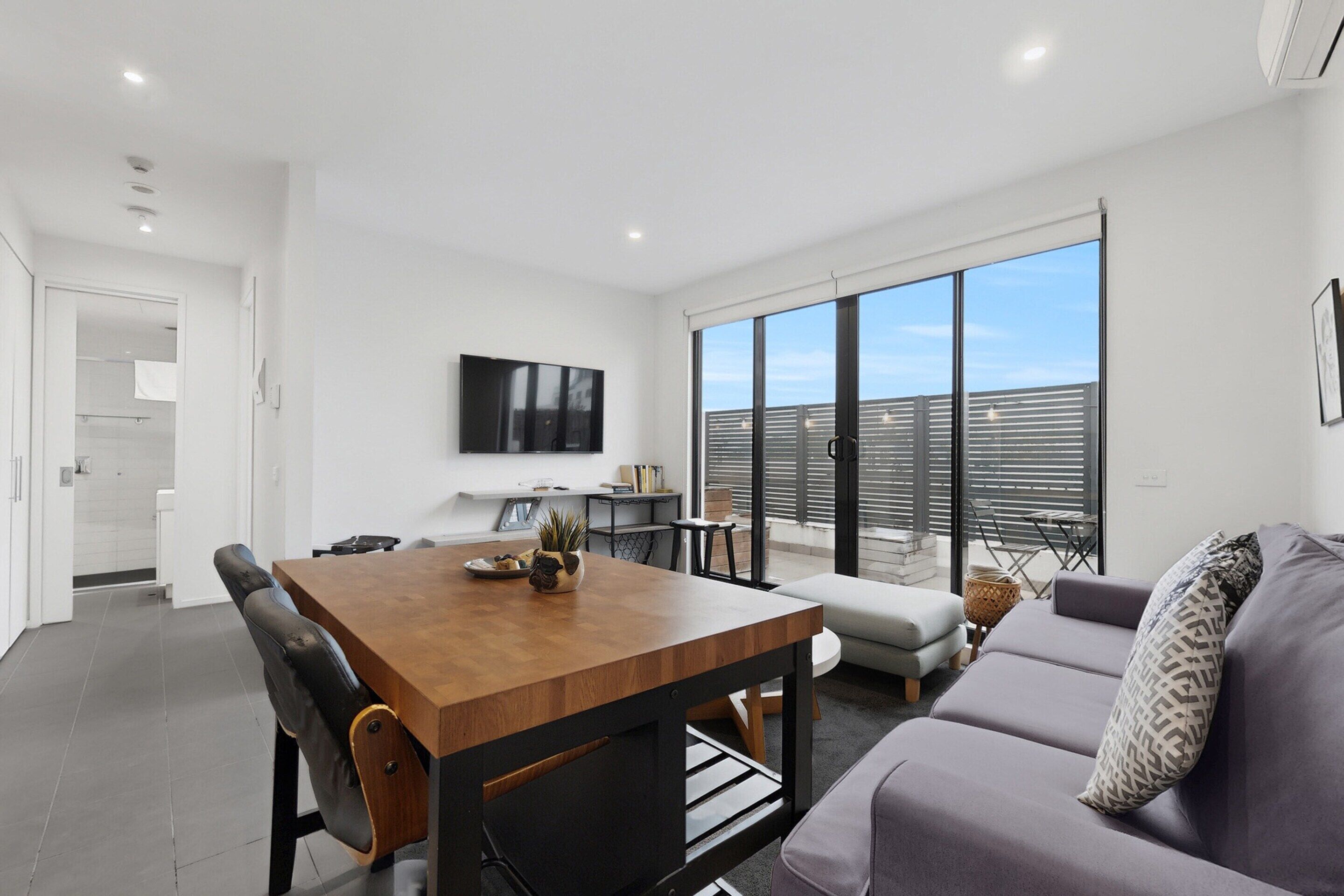 A modern living room with a wooden dining table, black chairs, a grey sofa, and a large window with blinds.