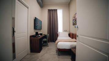 A compact hotel room with a bed, a desk, a chair, a window with curtains, and a wall-mounted air conditioner.