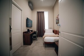 Une chambre d’hôtel de taille réduite, comprenant un lit, un bureau, une chaise, une fenêtre avec des rideaux et un climatiseur fixé au mur.