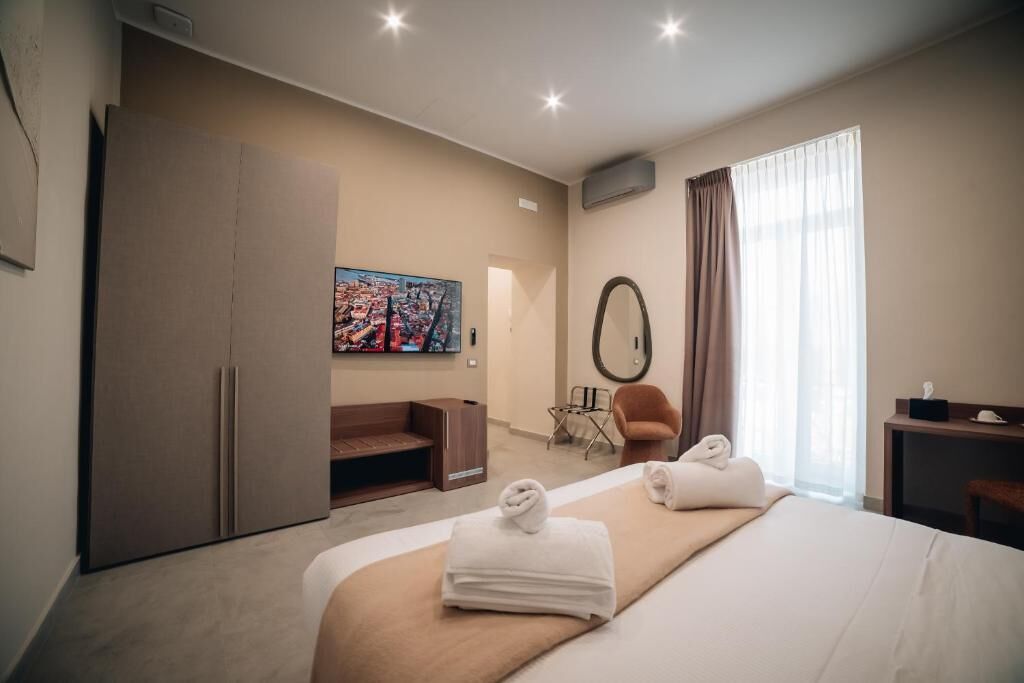 A hotel room with a bed, a desk, a chair, a wardrobe, a painting, and a window with curtains.