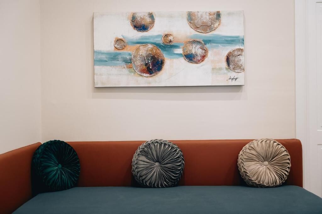A room with a painting on the wall, a brown sofa with three decorative pillows, and a teal-colored area rug.