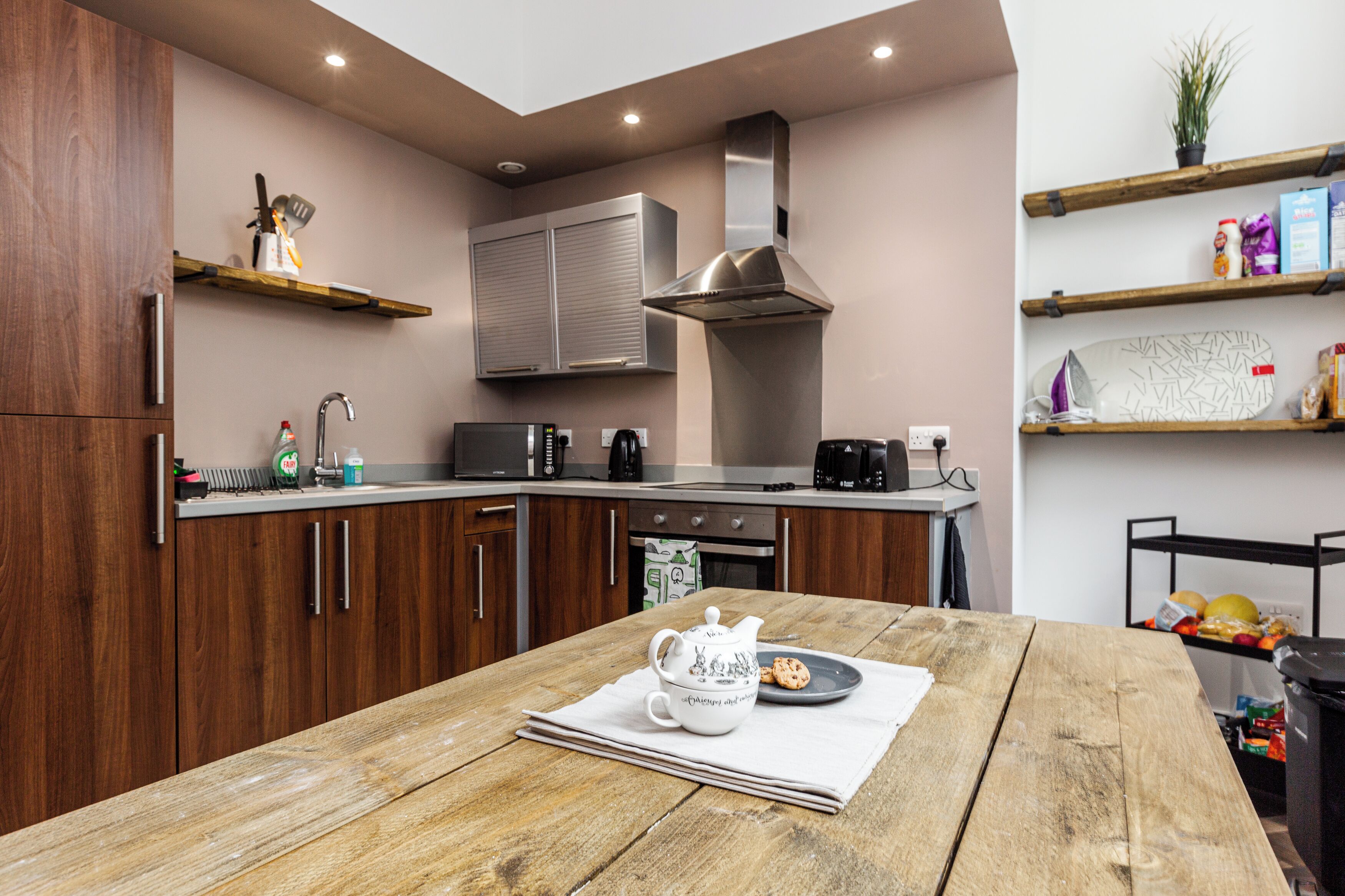 A modern kitchen with wooden cabinets, stainless steel appliances, and a wooden dining table with a teapot and a plate of cookies.