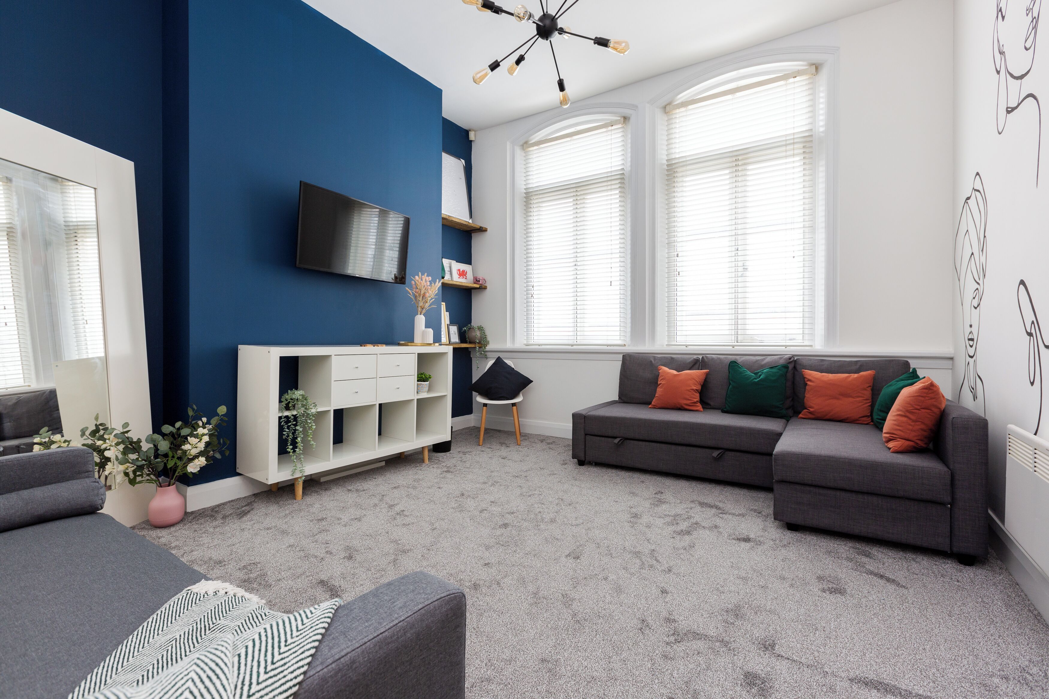 A modern living room with a blue accent wall, a grey sofa with colorful cushions, a white entertainment unit, and a large window with blinds.