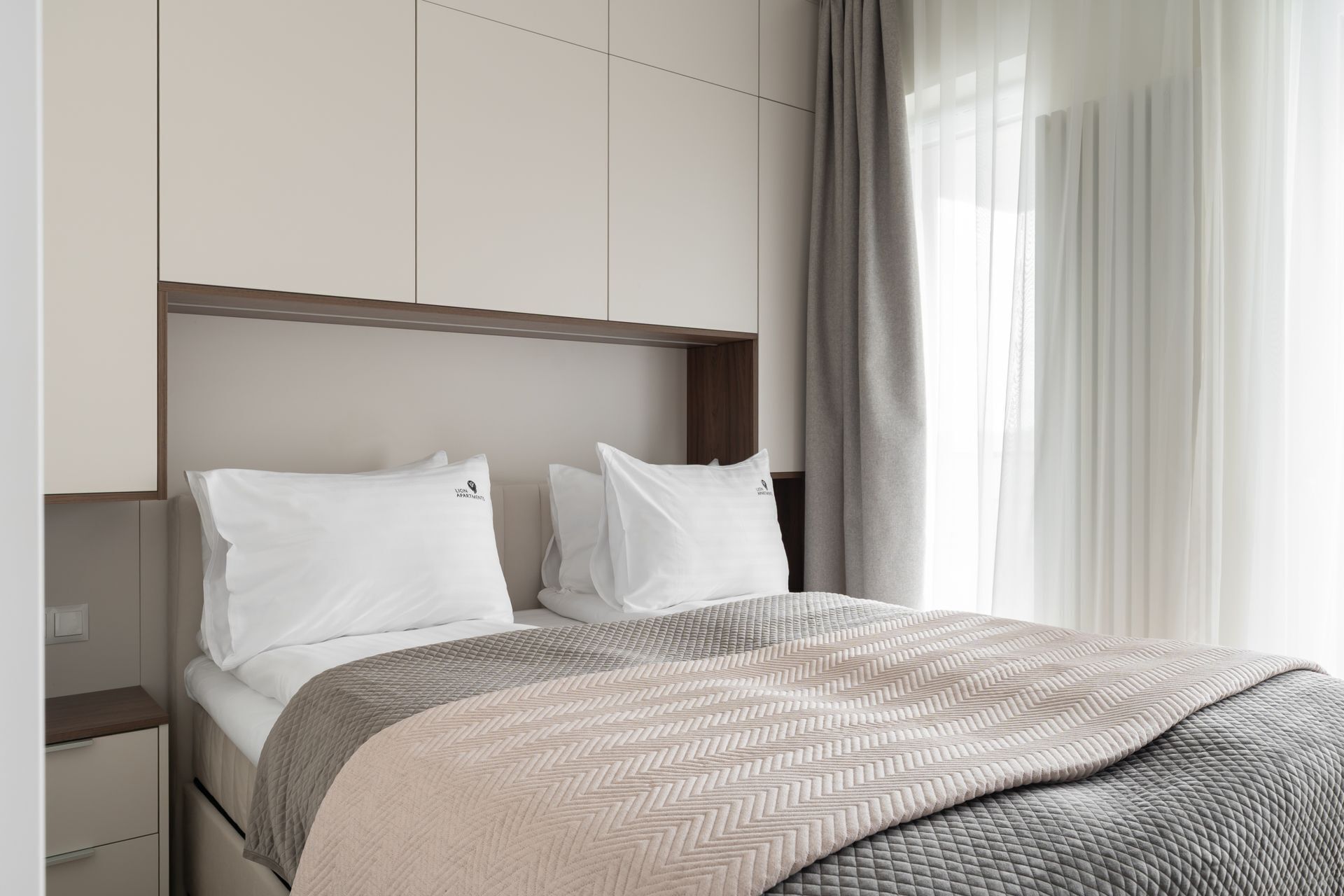 A hotel room with a bed, two pillows, a headboard, a bedside table, and a window with curtains.