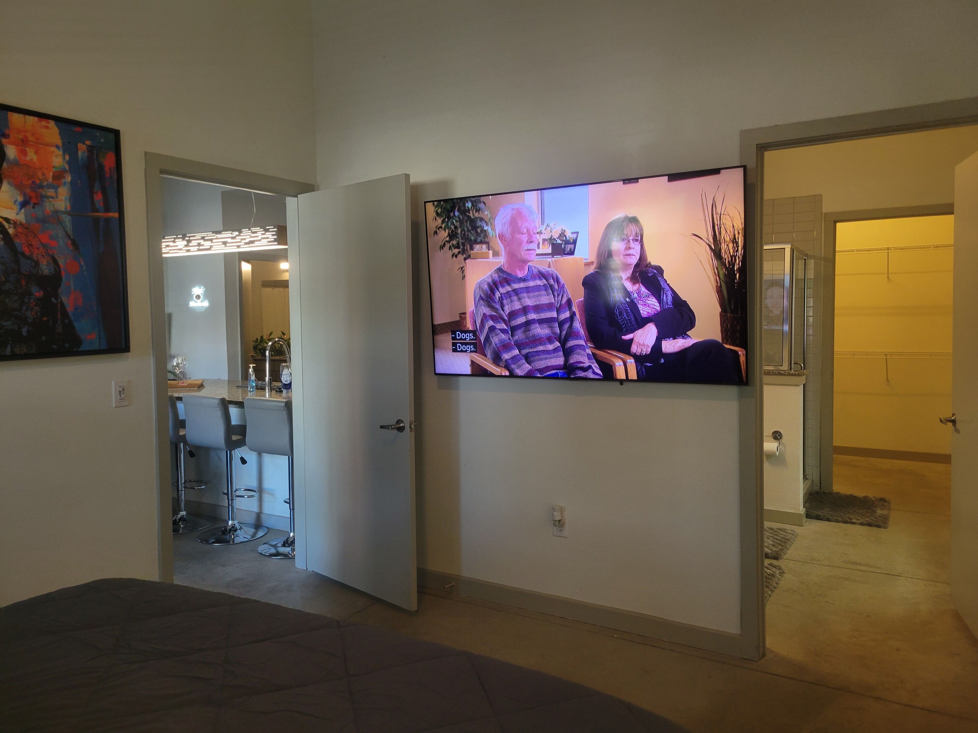 A hallway with a television displaying a scene, a dining area with chairs and a table, and a door leading to another room.