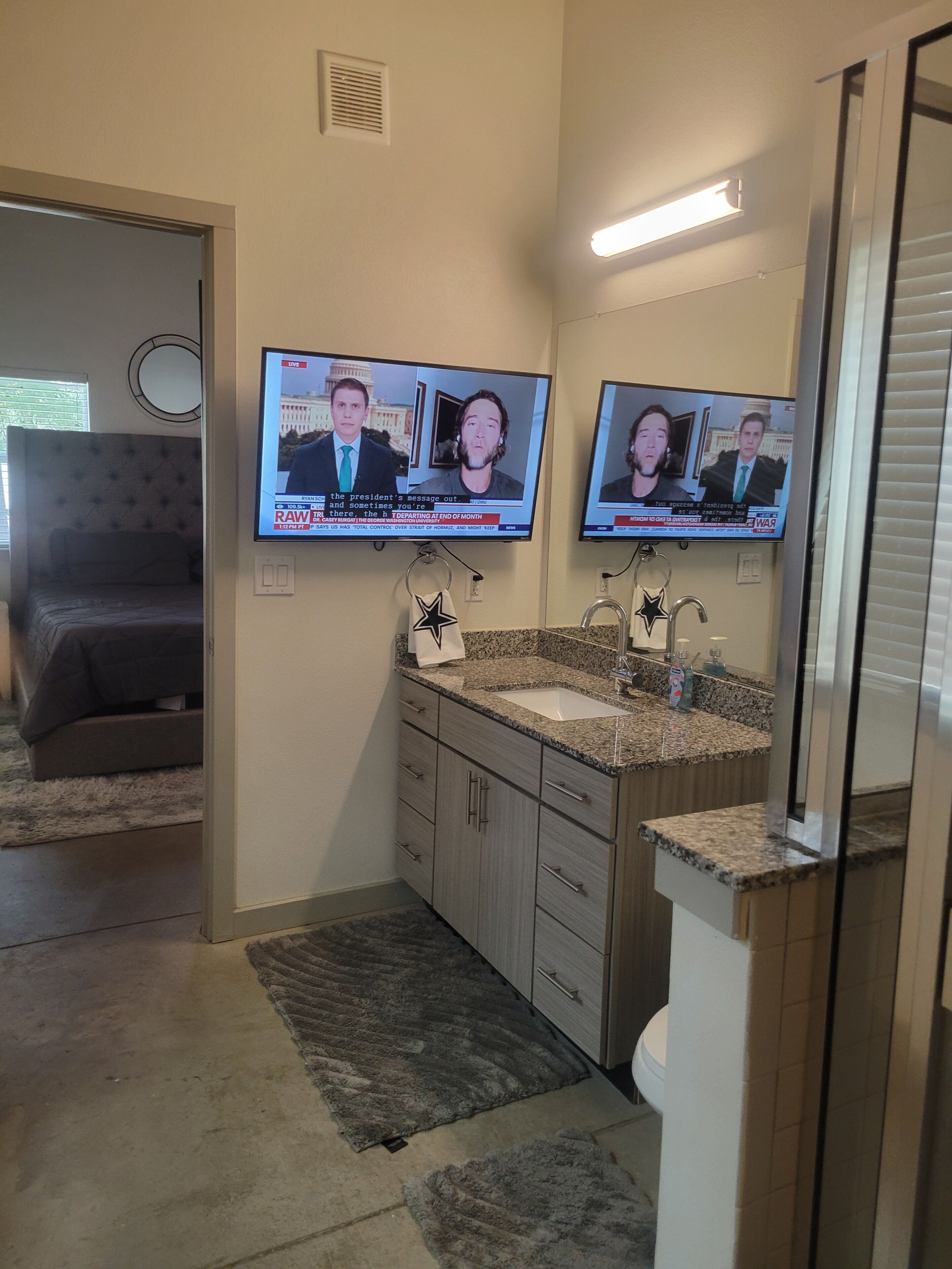 A bathroom with a double vanity, a shower, and a television mounted on the wall displaying news.
