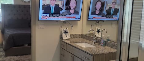 A bathroom with a double vanity, a shower, and a television mounted on the wall displaying news.
