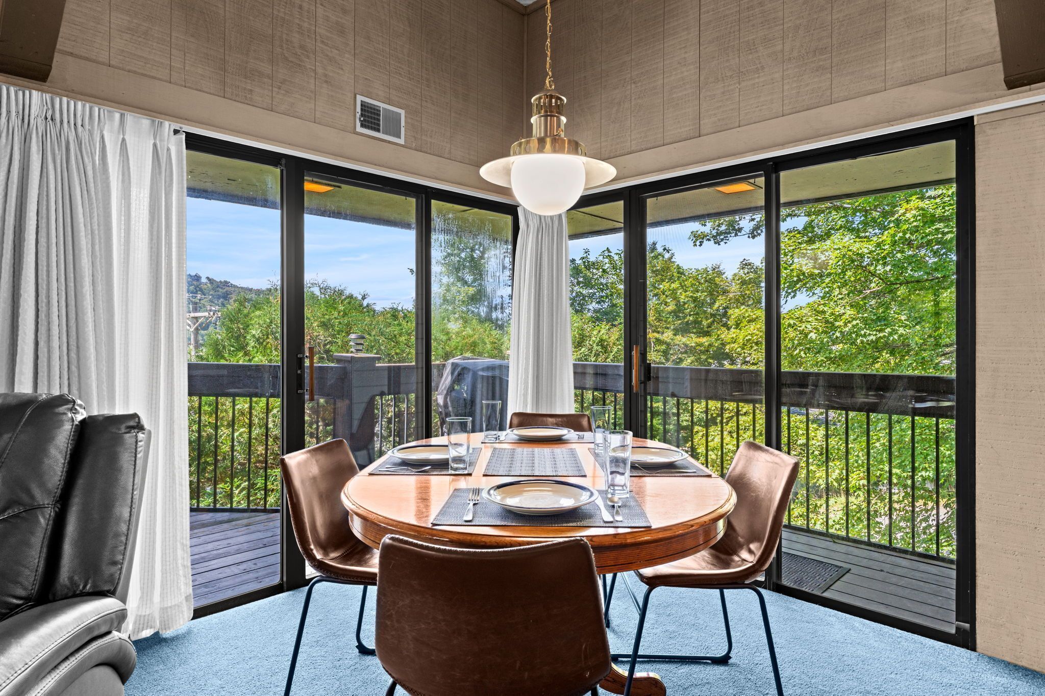 Un espace repas avec une table ronde dressée pour quatre personnes, entourée de fauteuils en cuir, de grandes fenêtres et offrant une vue sur la verdure.