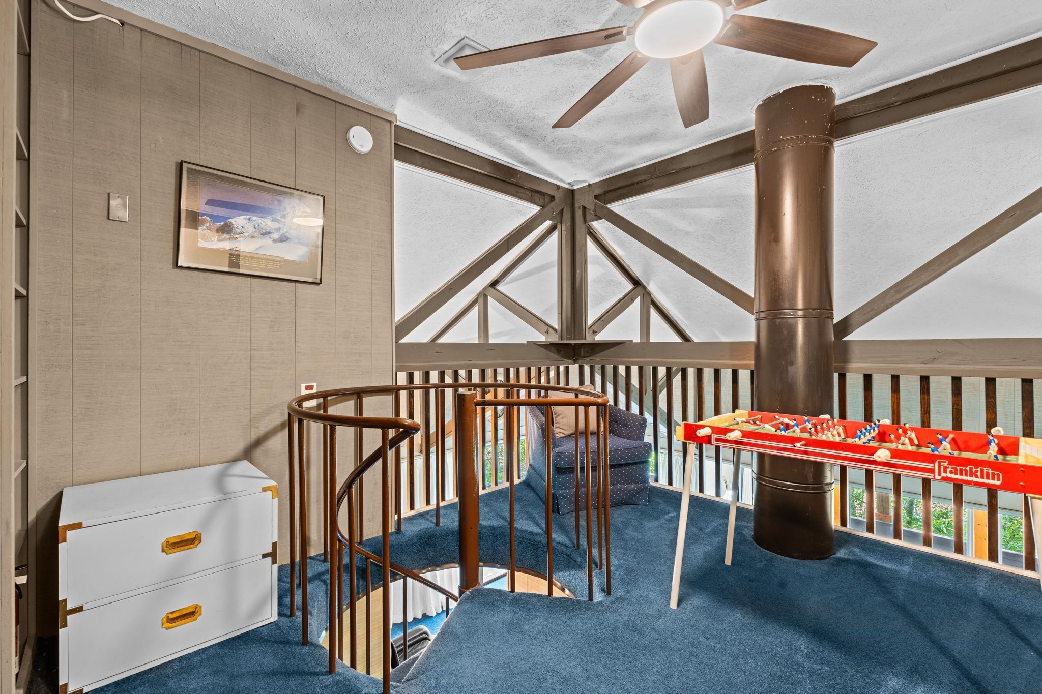 Une pièce avec un ventilateur de plafond, un tableau encadré, une commode blanche, un tapis bleu et une table de baby-foot.