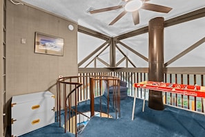 Une pièce avec un ventilateur de plafond, un tableau encadré, une commode blanche, un tapis bleu et une table de baby-foot.