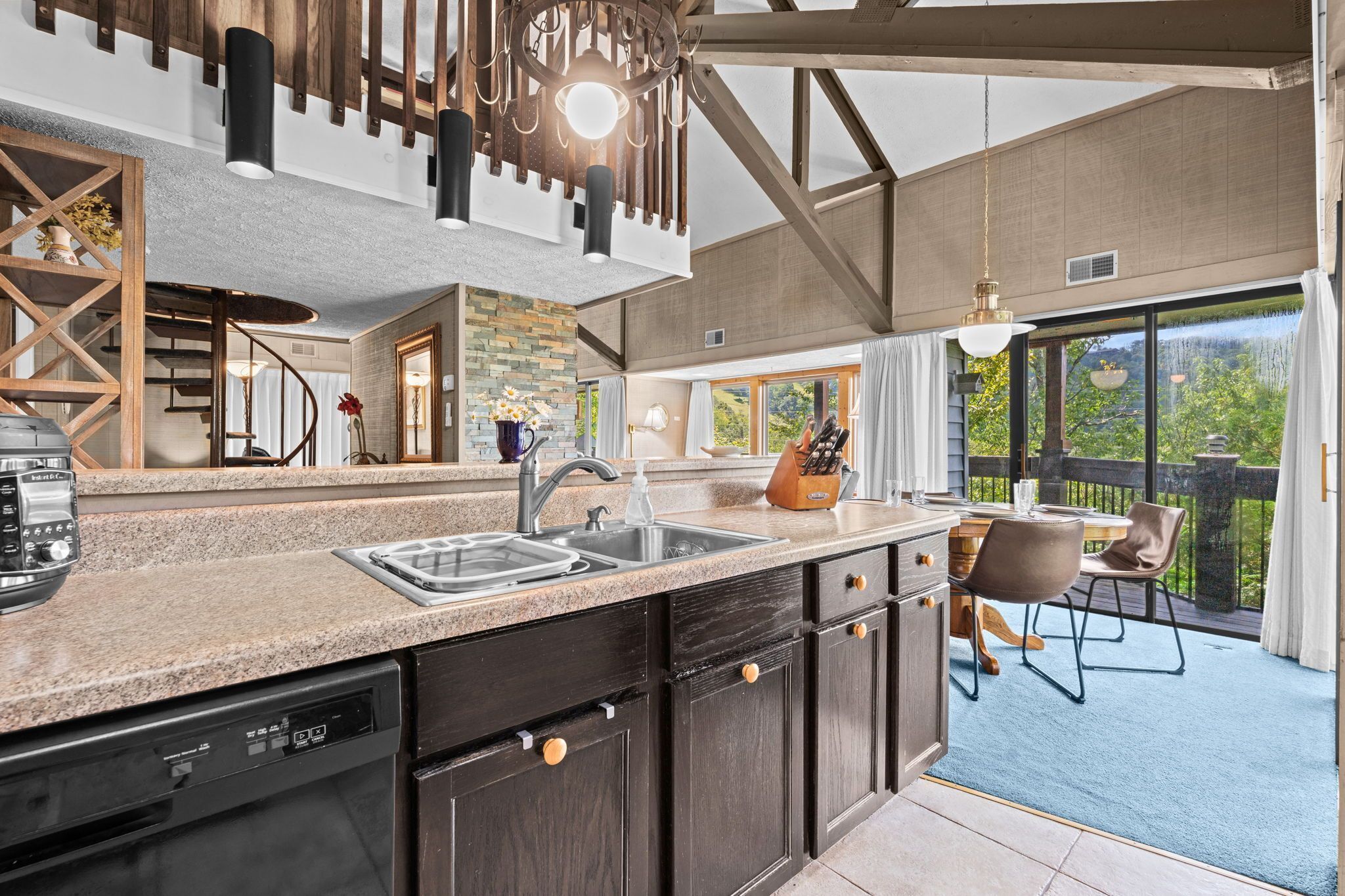A modern kitchen with granite countertops, a dishwasher, and a sink. There is a dining area with a table and chairs, and a view of the outdoors through large windows.