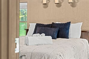 A bed with a white comforter, two rolled towels, and three pillows in shades of white and dark blue.