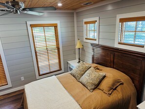 A bedroom with a bed, wooden headboard, two windows with blinds, a ceiling fan, and a bedside table with a lamp.