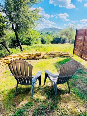Two Adirondack chairs facing a stone fire pit in a grassy outdoor area with a wooden fence and trees in the background.