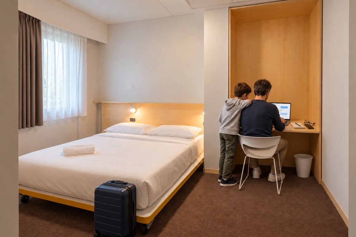 Una habitación de hotel con cama, escritorio, silla, maleta y un pequeño cubo de basura.