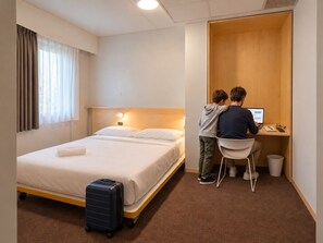 Una habitación de hotel con cama, escritorio, silla, maleta y un pequeño cubo de basura.
