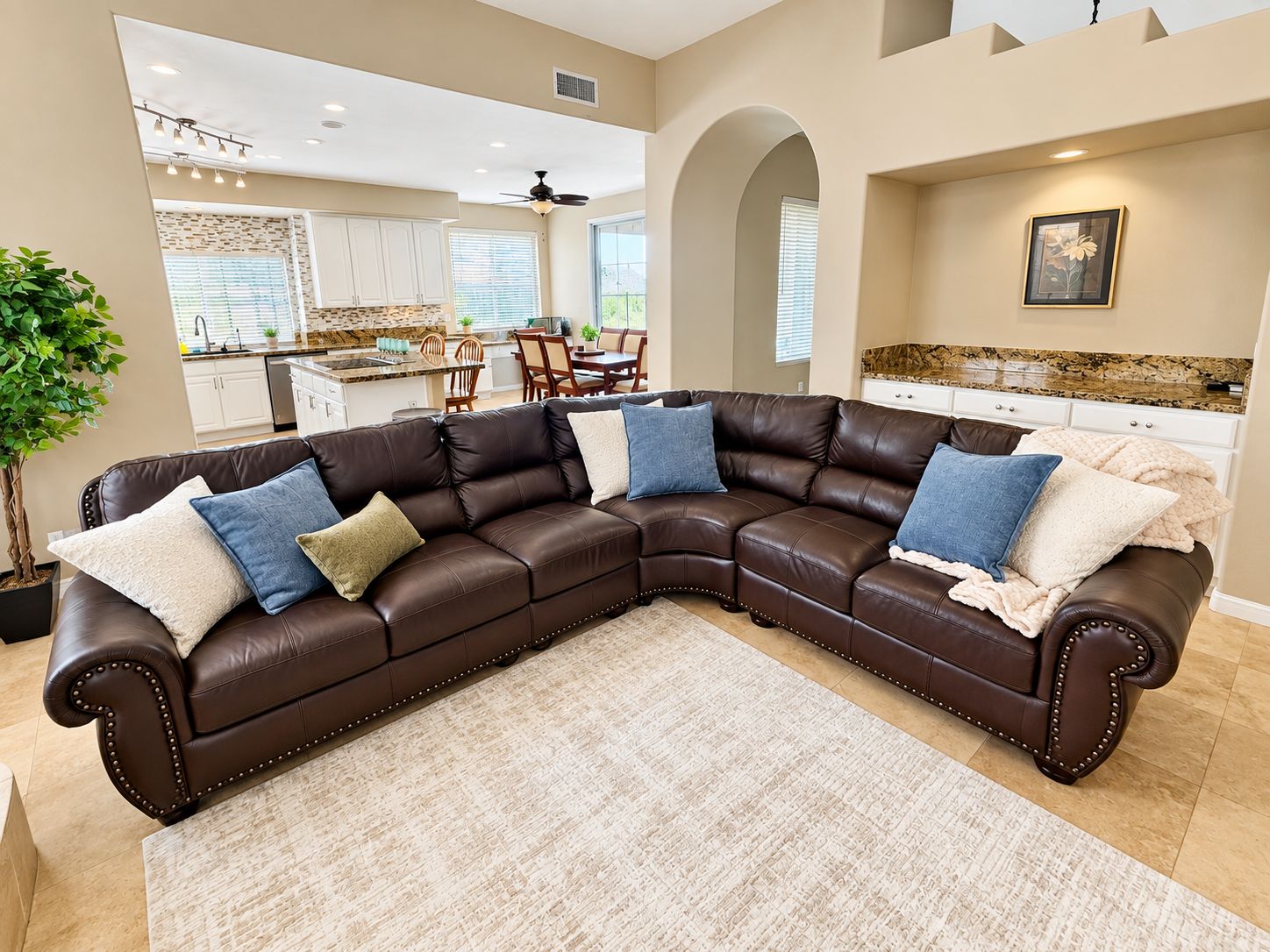 A spacious living room with a large L-shaped sectional sofa, a dining area, and a kitchen in the background.